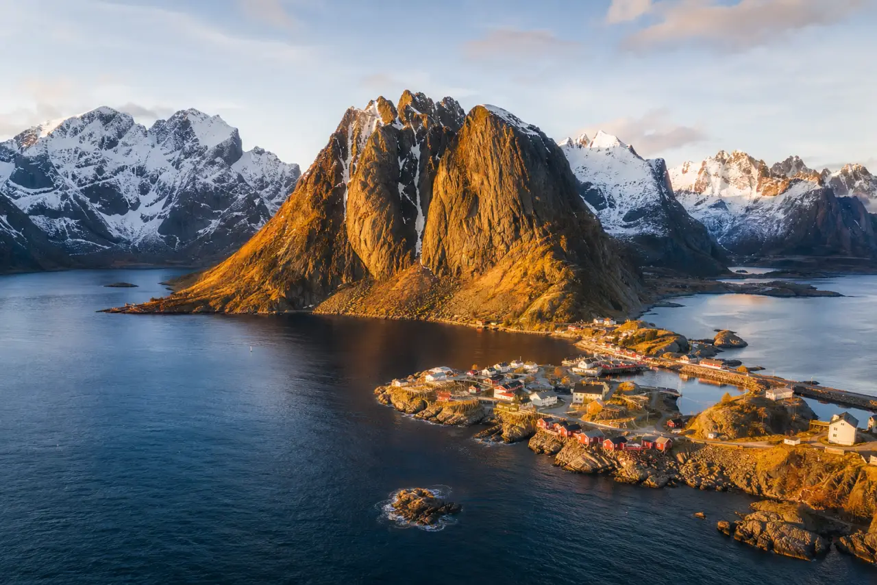 Aerial view of the Lofoten Islands in Norway at golden hour, with red fishing huts and mountain peaks reflected in the fjord